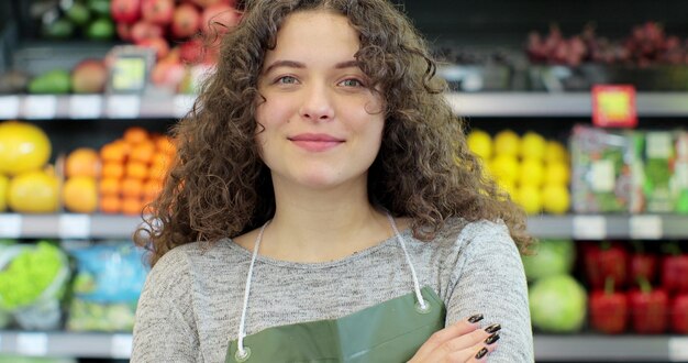 Joven trabajadora en un supermercado de la sección de frutas y verduras de pie sonriendo con los brazos cruzados Amable mujer mirando a la cámara en un mercado de tiendas