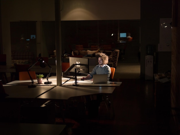 Foto joven trabajando en una computadora por la noche en una oficina oscura. el diseñador trabaja en la época posterior.