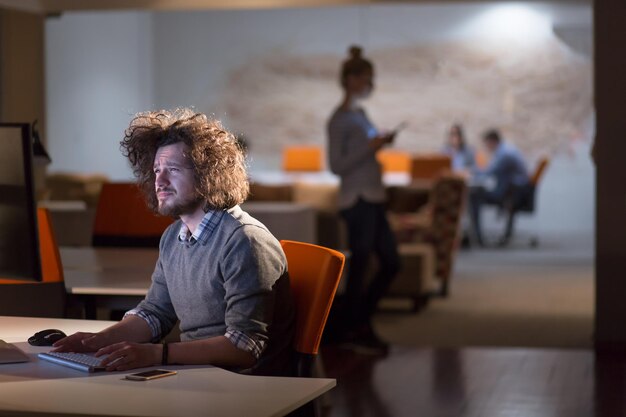 Joven trabajando en una computadora por la noche en una oficina oscura. El diseñador trabaja en la época posterior.