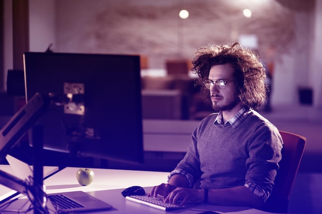 Joven trabajando en una computadora por la noche en una oficina oscura. El diseñador trabaja en la época posterior.