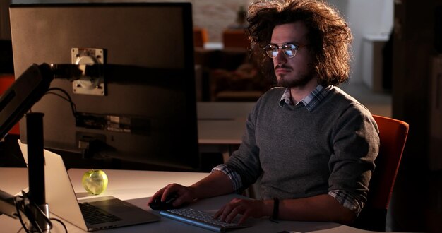 Joven trabajando en una computadora por la noche en una oficina oscura. El diseñador trabaja en la época posterior.