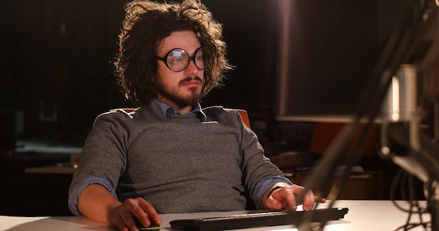 Joven trabajando en una computadora por la noche en una oficina oscura. El diseñador trabaja en la época posterior.