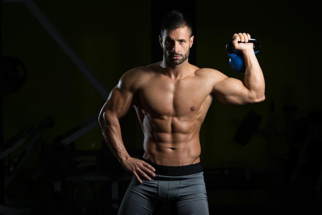 Joven trabajando con Kettle Bell en un gimnasio oscuro culturista haciendo ejercicio de peso pesado con pesas rusas