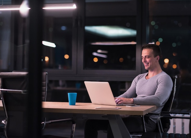 Joven trabajando en una laptop por la noche en una oficina oscura. El diseñador trabaja en la época posterior.