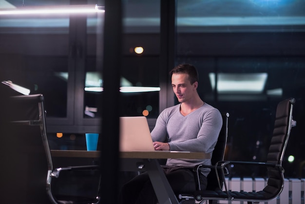 Joven trabajando en una laptop por la noche en una oficina oscura. El diseñador trabaja en la época posterior.