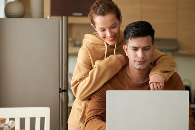 Joven trabajando en una laptop sentada en la mesa de la cocina con una mujer feliz abrazándolo por detrás