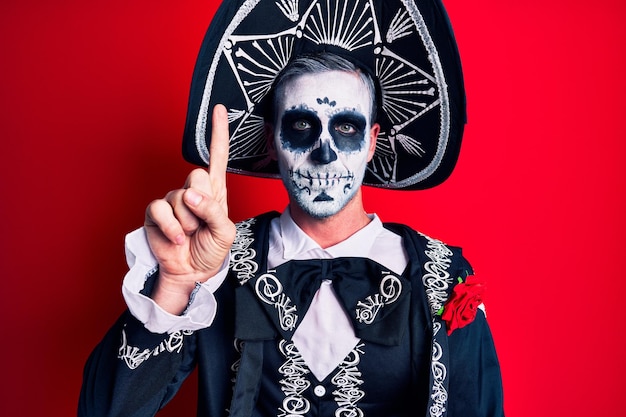 Joven con traje mexicano del día de los muertos sobre rojo que aparece y señala con el dedo número uno mientras sonríe confiado y feliz