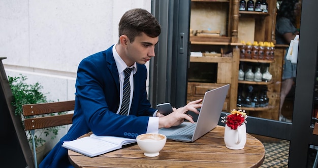 Joven con traje que sostiene un teléfono inteligente y navega por una computadora portátil mientras se sienta a la mesa en un acogedor café al aire libre en la calle de la ciudad