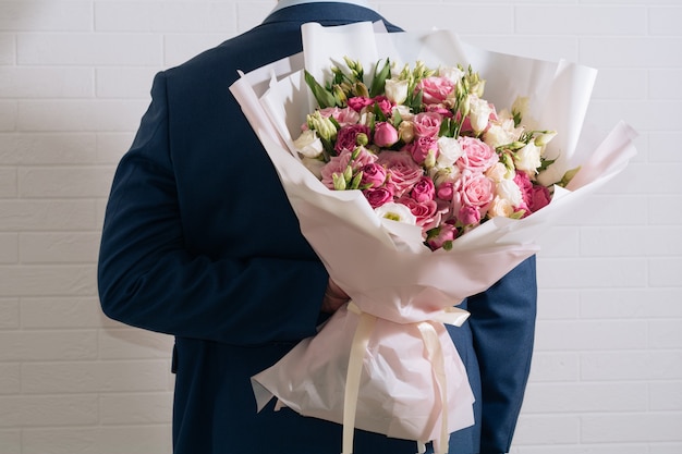 Joven en traje sostiene detrás de la espalda un gran ramo de rosas blancas y rosas eustoma