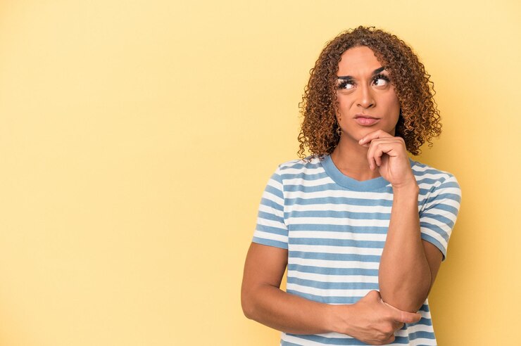 Foto joven transexual latina aislada de fondo amarillo mirando de lado con expresión dudosa y escéptica.