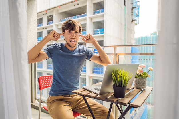 Joven tratando de trabajar en el balcón molesto por las obras de construcción fuera del concepto de ruido Contaminación del aire por el polvo del edificio