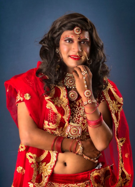 Joven travesti vestido con ropa de boda india de una novia con maquillaje nupcial y posando sobre fondo gris con aspecto de moda y glamuroso