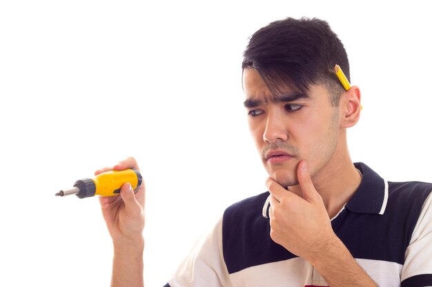 Joven triste con cabello negro en camiseta blanca y azul con lápiz amarillo detrás de la oreja sosteniendo un destornillador amarillo sobre fondo blanco en el estudio.