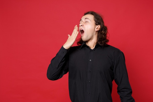 Joven triste hombre de pelo largo y rizado con camisa negra de pie posando aislado en un retrato de estudio de fondo de pared roja. Gente emociones sinceras concepto de estilo de vida. Simulacros de espacio de copia. Problema crisis depresión