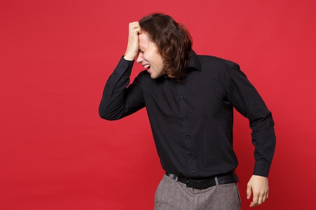 Joven triste hombre de pelo largo y rizado con camisa negra de pie posando aislado en un retrato de estudio de fondo de pared roja. Gente emociones sinceras concepto de estilo de vida. Simulacros de espacio de copia. Problema crisis depresión