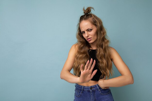 Joven triste molesta hermosa mujer rizada rubia encantadora con emociones sinceras vistiendo elegante top negro aislado sobre pared azul con espacio de copia y sin mostrar ningún gesto.