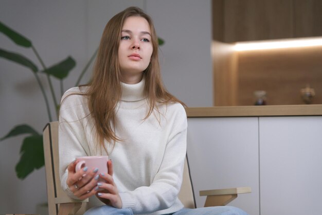 Una joven triste y pensativa está sentada en un cómodo y acogedor sillón en casa bebiendo té o café caliente en una taza con suéter en el frío día de invierno en la cocina