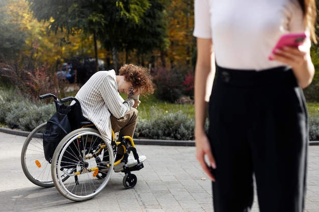 Un joven triste se sienta en una silla de ruedas sosteniendo su cabeza con la mano en la calle Una chica morena sostiene un teléfono inteligente en la mano