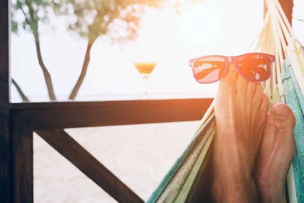 Joven tumbado en una hamaca en una playa soleada junto al mar. Gafas de sol en los pies