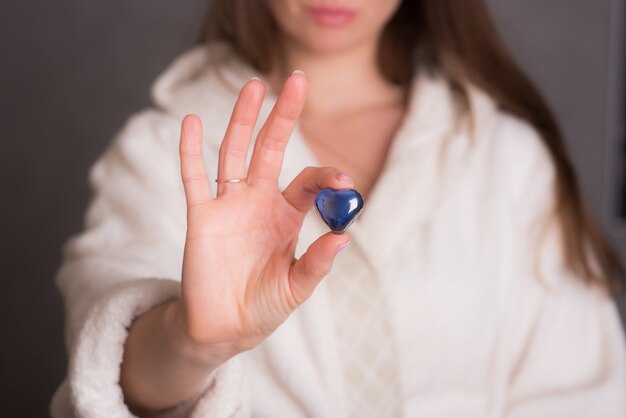 Una joven con una túnica blanca sostiene una piedra de cristal azul en la mano en forma de corazón. Piedra preciosa azul de gran plan en forma de corazón