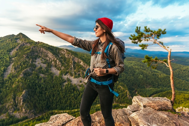 Una joven turista apuntando con su dedo a las montañas. Una turista con gafas de sol y una mochila caminando por las rocas en las montañas, sonríe y señala algo frente a ella. Viajes y senderismo