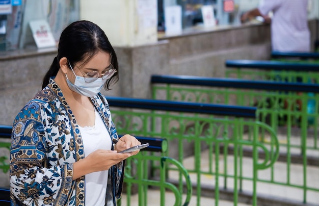 Joven turista asiática hipster con máscara protectora usando un teléfono inteligente en la estación de tren