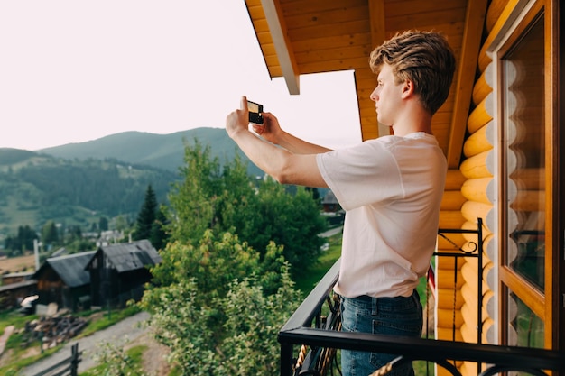 Un joven turista se para en el balcón de un apartamento en las montañas y toma una foto
