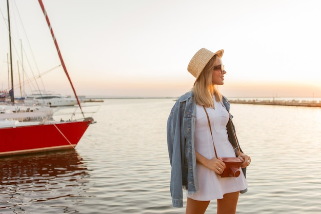 Joven turista con una cámara en la playa al amanecer vestida con sombrero, vestido blanco y gafas de sol