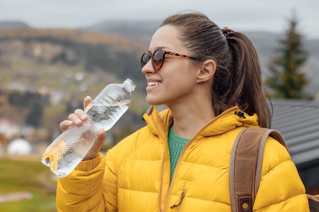 Joven turista con chaqueta amarilla bebe agua fresca en la naturaleza y el fondo de las montañas