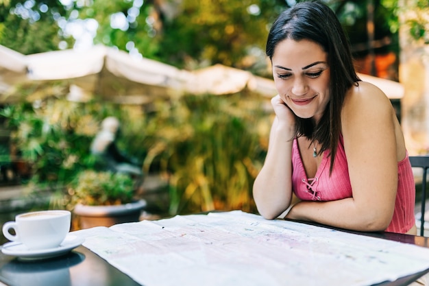 Foto joven turista hispano mirando un mapa mientras está sentado en un café. mujer de moda que usa un teléfono inteligente mientras viaja sola y busca nuevos lugares locales: vacaciones de verano y concepto de viajes por europa