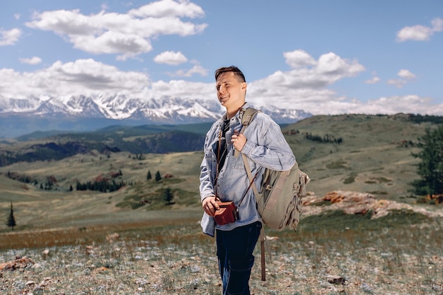 Un joven turista con una mochila está parado en un césped verde y sosteniendo una cámara en un estuche. Mirando hacia otro lado y observando la increíble vista a la montaña.