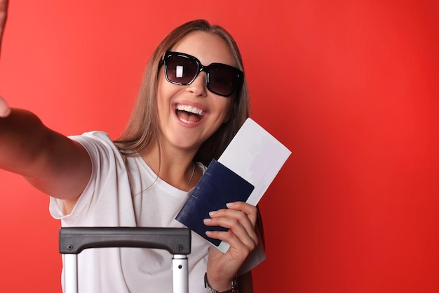 Joven turista con ropa informal de verano, con gafas de sol, maleta roja, pasaporte aislado de fondo rojo.