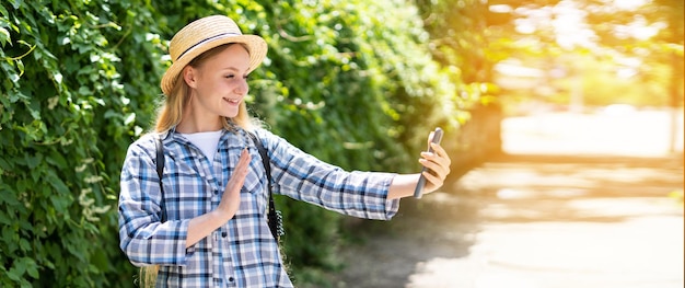 Joven turista rubia sonriendo feliz haciendo una videollamada usando un teléfono inteligente en la calle Fondo de arbustos y árboles verdes Concepto de vlog de viaje