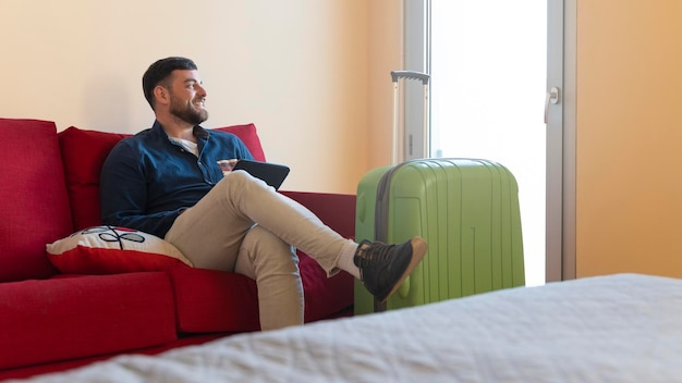 Joven turista con su tableta en una habitación de hotel sentada en un sofá mirando por la ventana