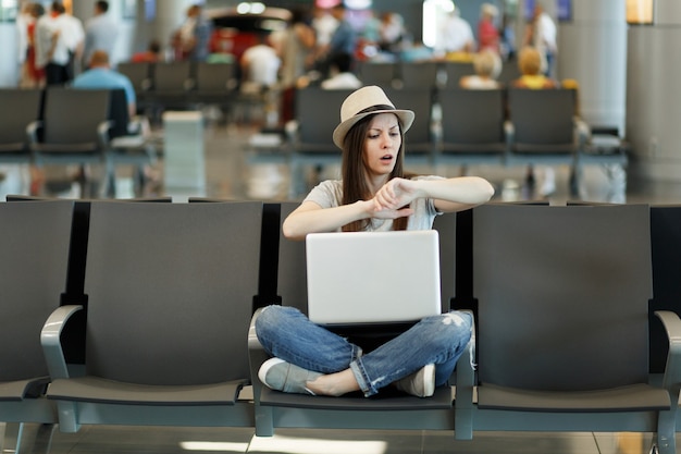 Foto joven turista viajero insatisfecho con laptop sentada con las piernas cruzadas, verificando el tiempo en el brazo, espera en el vestíbulo del aeropuerto