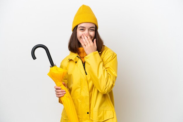 Joven ucraniana con abrigo impermeable y paraguas aislado de fondo blanco feliz y sonriente cubriendo la boca con la mano