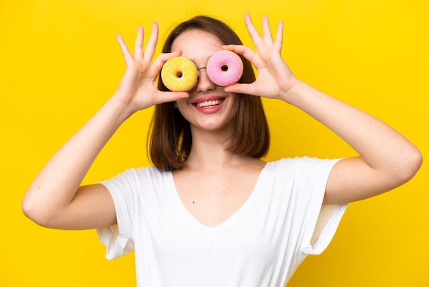 Joven ucraniana aislada de fondo amarillo sosteniendo donuts en los ojos