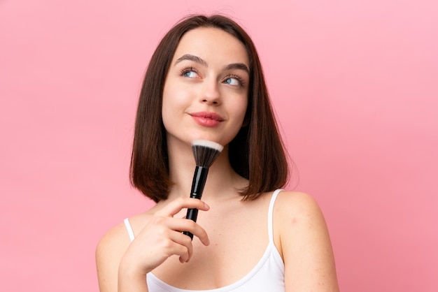 Joven ucraniana aislada de fondo rosa sosteniendo un pincel de maquillaje y pensando