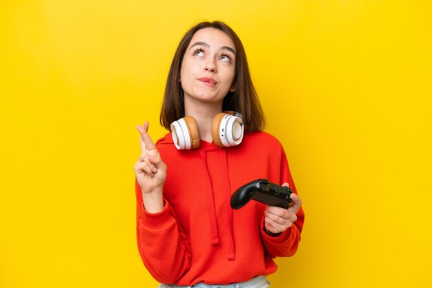 Joven ucraniana jugando con un controlador de videojuegos aislado en un fondo amarillo con los dedos cruzados y deseando lo mejor