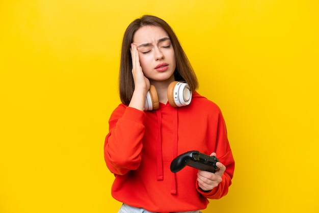 Joven ucraniana jugando con un controlador de videojuegos aislado de fondo amarillo con dolor de cabeza