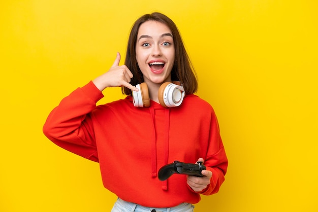 Joven ucraniana jugando con un controlador de videojuegos aislado en un fondo amarillo haciendo un gesto telefónico Llámame de nuevo