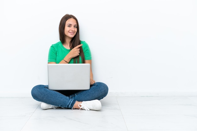 Joven ucraniana con una laptop sentada en el suelo apuntando hacia un lado para presentar un producto