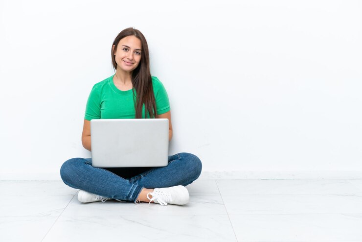Foto joven ucraniana con una laptop sentada en el suelo posando con los brazos en la cadera y sonriendo