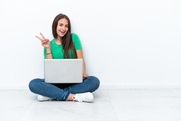 Joven ucraniana con una laptop sentada en el suelo sonriendo y mostrando el signo de la victoria