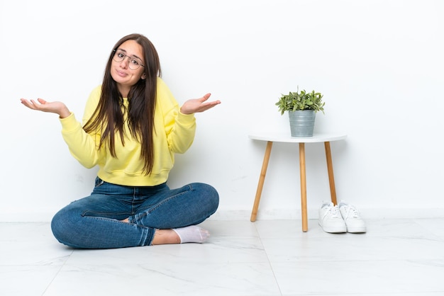 Foto joven ucraniana sentada en el piso de su casa aislada en una pared blanca que tiene dudas mientras levanta las manos