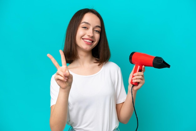 Joven ucraniano sosteniendo un secador de pelo aislado de fondo azul sonriendo y mostrando el signo de la victoria