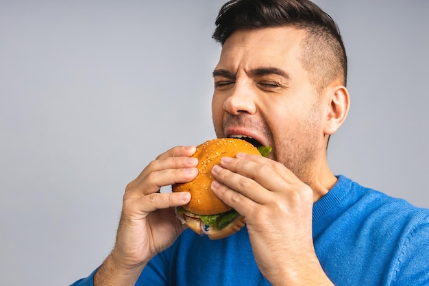 Joven ucraniano sosteniendo un trozo de hamburguesa El estudiante come comida rápida La hamburguesa no es un alimento útil Chico muy hambriento Concepto de dieta Aislado sobre fondo gris blanco