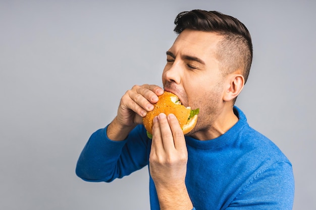 Joven ucraniano sosteniendo un trozo de hamburguesa El estudiante come comida rápida La hamburguesa no es un alimento útil Chico muy hambriento Concepto de dieta Aislado sobre fondo gris blanco