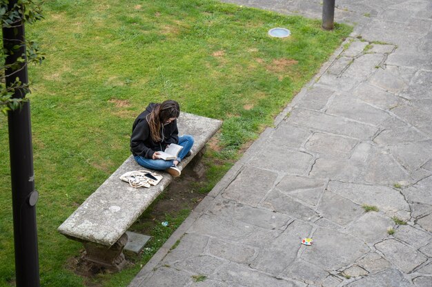 Joven universitaria leyendo un libro sentada en un banco de piedra en su descanso rodeada de una hermosa hierba verde