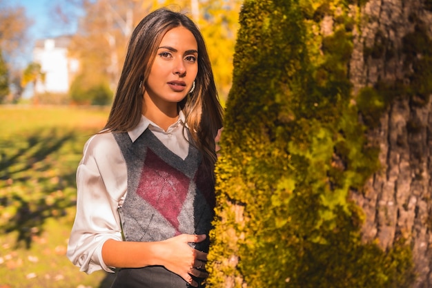 Una joven universitaria morena caucásica, en un parque junto a un árbol una tarde de otoño